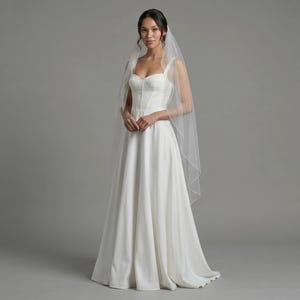 May include: A floor-length white wedding dress featuring a structured corset-style bodice with a sweetheart neckline and thick straps, paired with a sheer, lightweight tulle veil that drapes over the shoulders and flows down the back.