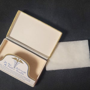 May include: An open white box with gold trim and a blue and gold medical symbol. The box contains a white cotton pad. The box has the text "In the Interest of Public Health" printed on it.