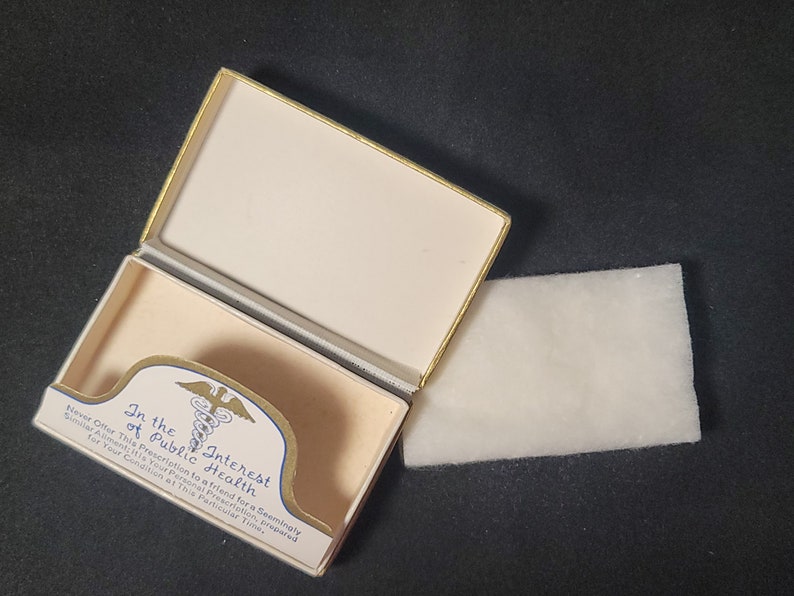 May include: An open white box with gold trim and a blue and gold medical symbol. The box contains a white cotton pad. The box has the text "In the Interest of Public Health" printed on it.