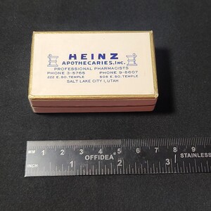 May include: A white box with gold trim and blue lettering. The box reads "Heinz Apothecaries, Inc." and includes the company's phone numbers and address: "Professional Pharmacists, Phone 3-5765, 222 E. So. Temple, Phone 9-6607, 508 E. So. Temple, Salt Lake City 1, Utah".