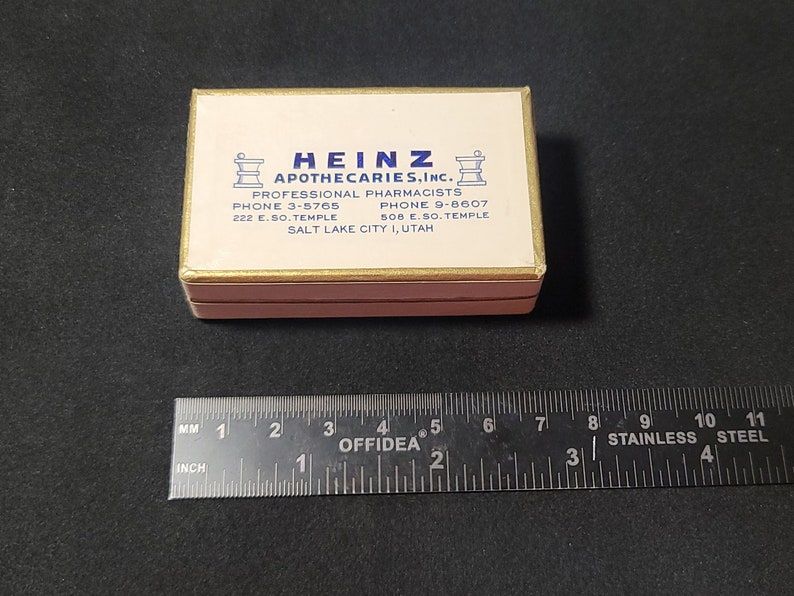 May include: A white box with gold trim and blue lettering. The box reads "Heinz Apothecaries, Inc." and includes the company's phone numbers and address: "Professional Pharmacists, Phone 3-5765, 222 E. So. Temple, Phone 9-6607, 508 E. So. Temple, Salt Lake City 1, Utah".