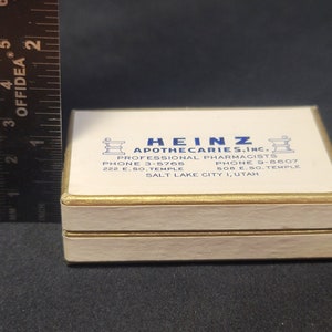 May include: A white box with gold trim and the text "HEINZ APOTHECARIES, INC." in blue lettering. The box also includes the text "PROFESSIONAL PHARMACISTS", "PHONE 3-5765", "PHONE 9-8607", "222 E. SO. TEMPLE", "508 E. SO. TEMPLE", and "SALT LAKE CITY 1, UTAH".