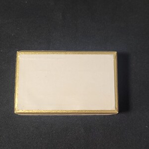 May include: A small, rectangular box with a white interior and a gold border. The box is closed.