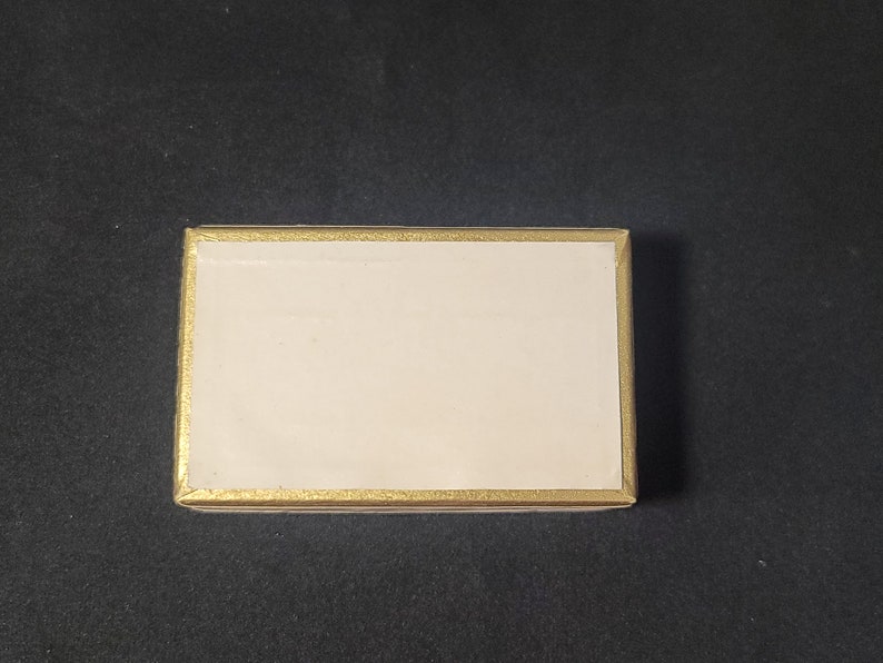 May include: A small, rectangular box with a white interior and a gold border. The box is closed.