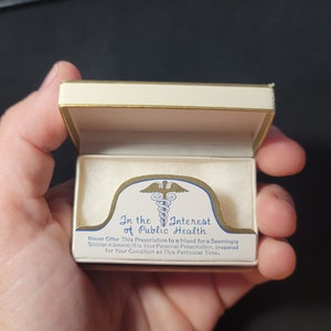 May include: A white box with a gold trim and a gold and blue medical symbol. The box has the text "In the Interest of Public Health" and "Never Offer This Prescription to a friend for a Seemingly Similar Ailment: It is Your Personal Prescription, prepared for Your Condition at This Particular Time."