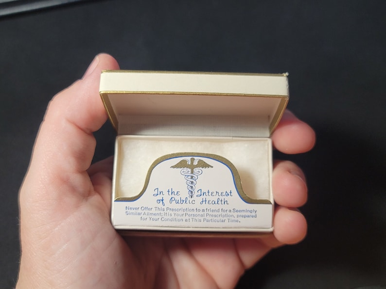 May include: A white box with a gold trim and a gold and blue medical symbol. The box has the text "In the Interest of Public Health" and "Never Offer This Prescription to a friend for a Seemingly Similar Ailment: It is Your Personal Prescription, prepared for Your Condition at This Particular Time."