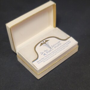 May include: A white box with a gold trim, open to reveal a white card with a blue and gold medical symbol and the text "In the Interest of Public Health. Never Offer This Prescription to a friend for a Seemingly Similar Ailment: It is Your Personal Prescription, prepared for Your Condition at This Particular Time."