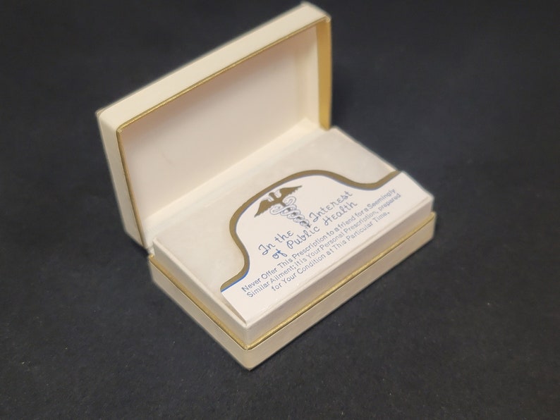 May include: A white box with a gold trim, open to reveal a white card with a blue and gold medical symbol and the text "In the Interest of Public Health. Never Offer This Prescription to a friend for a Seemingly Similar Ailment: It is Your Personal Prescription, prepared for Your Condition at This Particular Time."