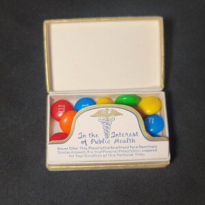 May include: A small, white box with a gold trim, open to reveal a small card with the text "In the Interest of Public Health" and a medical symbol. The box is filled with colorful M&M's candies.