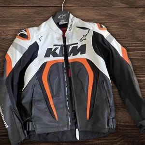 Könnte beinhalten: Eine schwarze, weiße und orangefarbene Motorradjacke mit dem KTM-Logo. Die Jacke hat einen durchgehenden Reißverschluss, mehrere Taschen und gepolsterte Schultern. Die Jacke hängt an einem Kleiderbügel.