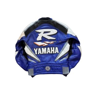 K&ouml;nnte beinhalten: Blaue und wei&szlig;e Motorradjacke aus Leder mit dem Yamaha-Logo auf dem R&uuml;cken. Die Jacke hat einen schwarzen Kragen und wei&szlig;e Akzente an den &Auml;rmeln. Auf der R&uuml;ckseite der Jacke befindet sich ein gro&szlig;es wei&szlig;es "R" und das Wort "YAMAHA".