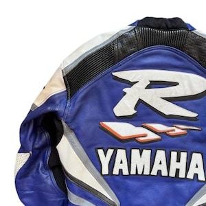 K&ouml;nnte beinhalten: Blaue, wei&szlig;e und schwarze Motorradjacke aus Leder mit dem Yamaha-Logo. Die Jacke hat ein gro&szlig;es wei&szlig;es "R" und "R6" Design auf dem R&uuml;cken mit orangefarbenen Akzenten. Die Jacke hat einen gepolsterten Kragen und Schulterdetails.