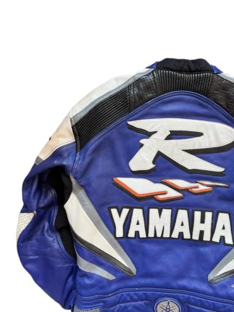 K&ouml;nnte beinhalten: Blaue, wei&szlig;e und schwarze Motorradjacke aus Leder mit dem Yamaha-Logo. Die Jacke hat ein gro&szlig;es wei&szlig;es "R" und "R6" Design auf dem R&uuml;cken mit orangefarbenen Akzenten. Die Jacke hat einen gepolsterten Kragen und Schulterdetails.