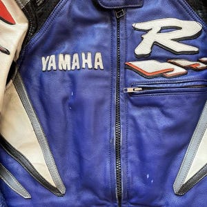 K&ouml;nnte beinhalten: Nahaufnahme einer Motorradjacke aus blauem und wei&szlig;em Leder. Die Jacke hat das Yamaha-Logo in Wei&szlig; sowie den Markennamen Lookwell. Die Jacke hat einen Rei&szlig;verschluss und wei&szlig;e und rote Akzente.