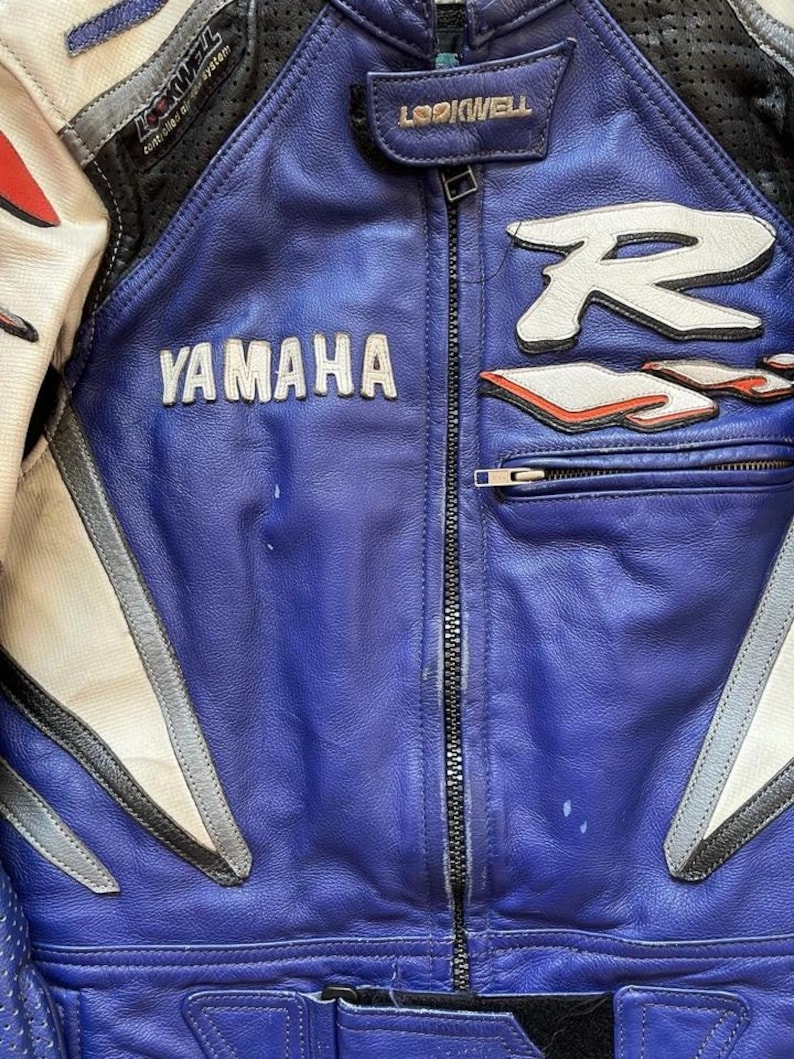 K&ouml;nnte beinhalten: Nahaufnahme einer Motorradjacke aus blauem und wei&szlig;em Leder. Die Jacke hat das Yamaha-Logo in Wei&szlig; sowie den Markennamen Lookwell. Die Jacke hat einen Rei&szlig;verschluss und wei&szlig;e und rote Akzente.