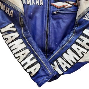 K&ouml;nnte beinhalten: Blaue Yamaha Motorrad-Rennjacke mit wei&szlig;en und roten Details. Das Yamaha-Logo in Wei&szlig; ist auf den &Auml;rmeln und der Brust zu sehen. Die Jacke hat einen Rei&szlig;verschluss und eine Brusttasche. Aus Leder.
