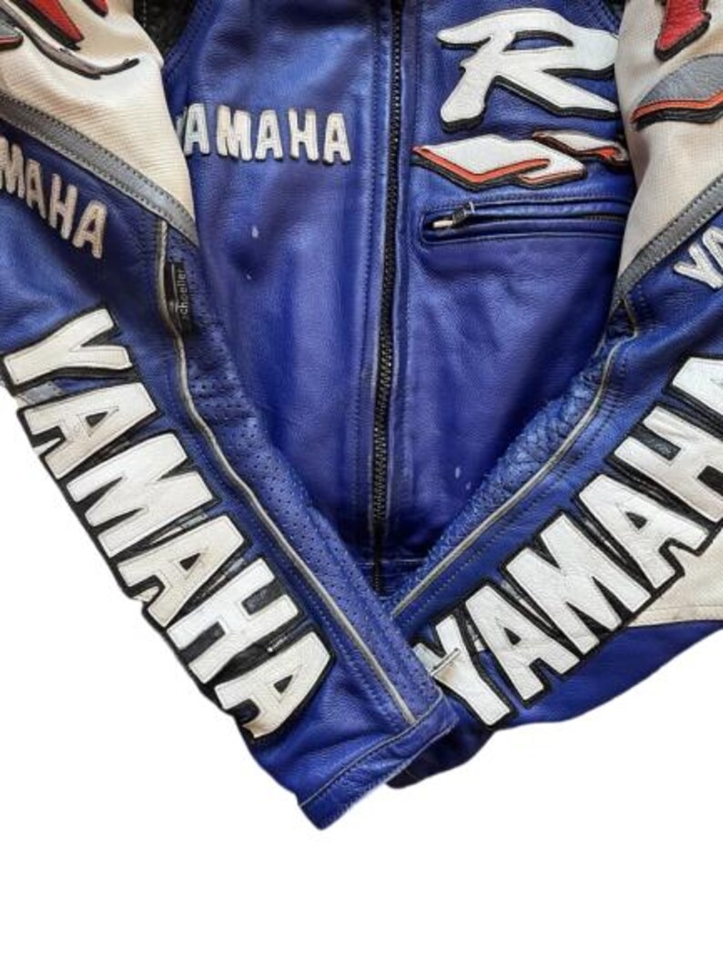 K&ouml;nnte beinhalten: Blaue Yamaha Motorrad-Rennjacke mit wei&szlig;en und roten Details. Das Yamaha-Logo in Wei&szlig; ist auf den &Auml;rmeln und der Brust zu sehen. Die Jacke hat einen Rei&szlig;verschluss und eine Brusttasche. Aus Leder.