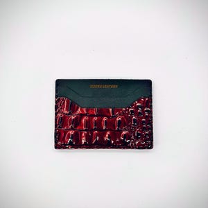May include: A rectangular card holder with a glossy, textured, burgundy crocodile-patterned leather front. The top section is dark green leather with the text "BLICKS LEATHER" in gold lettering.