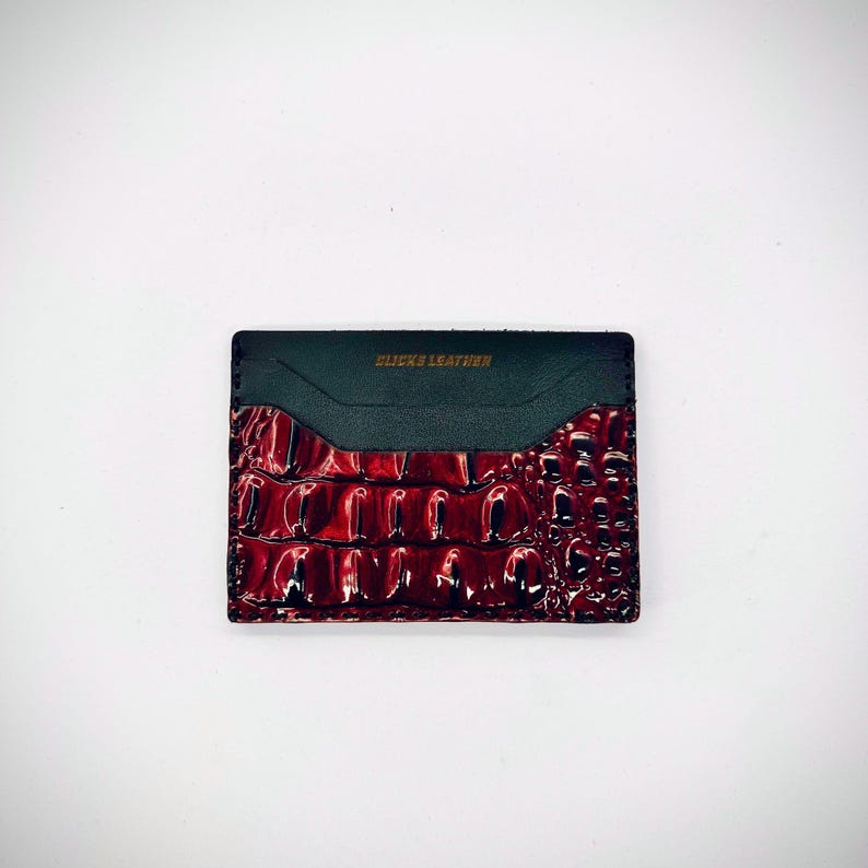 May include: A rectangular card holder with a glossy, textured, burgundy crocodile-patterned leather front. The top section is dark green leather with the text "BLICKS LEATHER" in gold lettering.