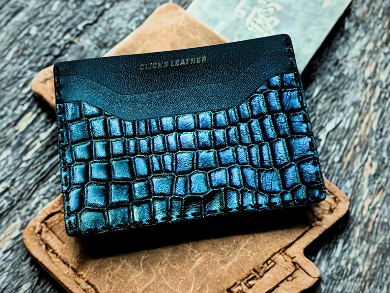 以下が含まれることがあります： クロコダイル柄の青と黒のレザーカードホルダー。カードホルダーの上部には「CLICKS LEATHER」の文字がエンボス加工されています。カードホルダーは茶色の革の表面に置かれています。