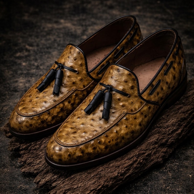 May include: A pair of brown leather loafers with a textured pattern and black tassels. The shoes have a classic design with a rounded toe and a dark sole. The loafers are displayed on a wooden surface.