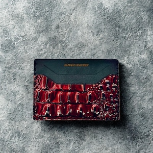 May include: A rectangular cardholder with a dark teal top and a textured, glossy, burgundy-coloured bottom. The text "CLICKS LEATHER" is printed in gold on the top. The cardholder is likely made of leather.