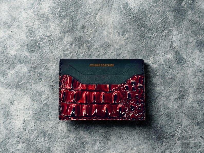 May include: A rectangular cardholder with a dark teal top and a textured, glossy, burgundy-coloured bottom. The text "CLICKS LEATHER" is printed in gold on the top. The cardholder is likely made of leather.