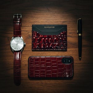 May include: A coordinated set of accessories in burgundy crocodile-effect leather, including a wristwatch, cardholder, phone case, and pen. The watch has a silver face and the cardholder is labelled "SLICKS LEATHER".