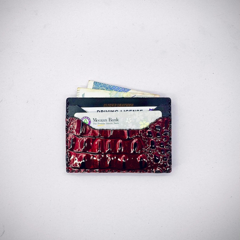 May include: A burgundy leather cardholder with a crocodile skin texture. The cardholder contains a driving licence, a bank card, and some banknotes. The cardholder is black on the inside and has the text "SLICK LEATHER" on the top.
