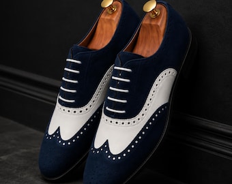 May include: A pair of navy blue suede oxford shoes featuring white leather wingtip detailing, white laces, and perforated brogue accents. Each shoe includes a wooden shoe tree with a gold-toned knob inserted into the opening for shape retention.