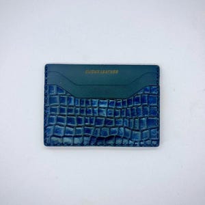 以下が含まれることがあります： クロコダイル柄の青い革製カードケース。上部は無地の濃いティール色で、下部はテクスチャード加工されたエンボス加工のデザインです。「CLICKS LEATHER」の文字が金色の文字でエンボス加工されています。