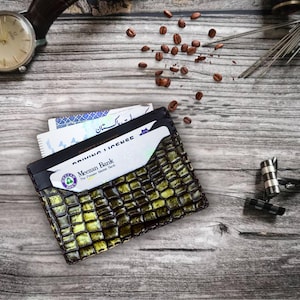 May include: A textured, olive green and black leather wallet with cards and a driver's license peeking out. A brown leather watch, coffee beans, and cufflinks are also visible on a wooden surface.