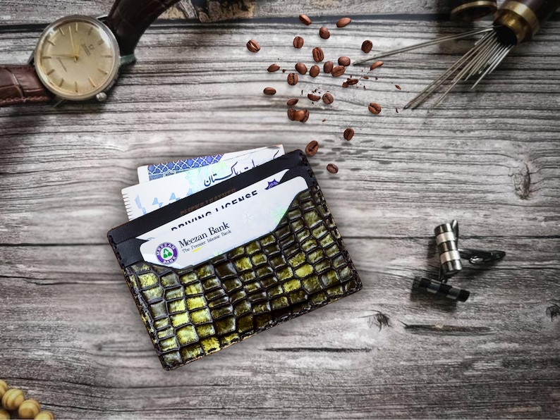 May include: A textured, olive green and black leather wallet with cards and a driver's license peeking out. A brown leather watch, coffee beans, and cufflinks are also visible on a wooden surface.