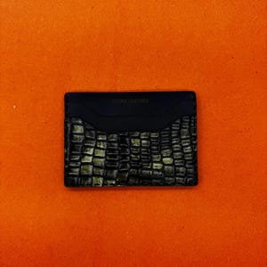 May include: Black leather card holder with a crocodile skin pattern. The top section is smooth, and the bottom has a textured, embossed design. The text "CLICKS LEATHER" is embossed on the top.