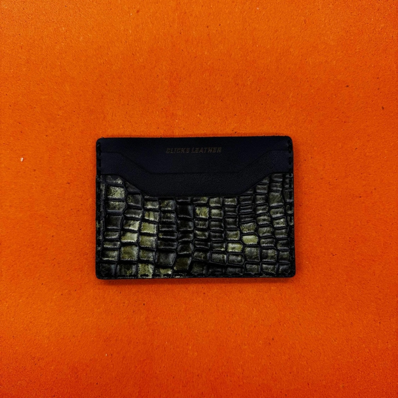 May include: Black leather card holder with a crocodile skin pattern. The top section is smooth, and the bottom has a textured, embossed design. The text "CLICKS LEATHER" is embossed on the top.