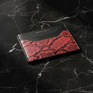 May include: A black and red cardholder with a snakeskin pattern. The top section is black, with the text "CHICKS LEATHER" printed in gold. The cardholder is rectangular and rests on a black marble surface.