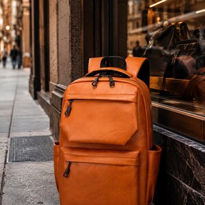 May include: A brown leather backpack with multiple zippered compartments and a top handle. The bag is standing upright on a sidewalk in front of a store window, with a blurred city street in the background.