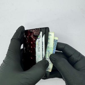 May include: A black wallet with a textured, dark red front panel, held by gloved hands. The wallet contains a driver's license, a bank card, and several banknotes. The text "DRIVING LICENSE" is visible.