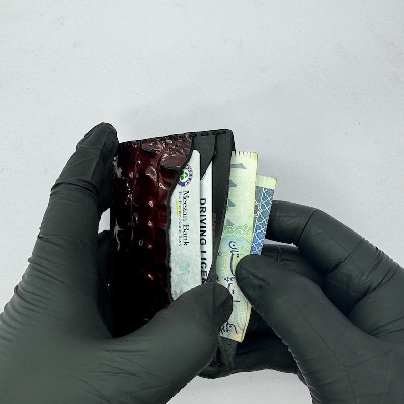 May include: A black wallet with a textured, dark red front panel, held by gloved hands. The wallet contains a driver's license, a bank card, and several banknotes. The text "DRIVING LICENSE" is visible.