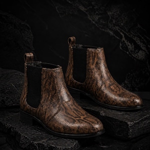 May include: A pair of brown ankle boots with a snakeskin pattern. The boots have black elastic side panels and pull tabs. They are displayed on a dark, textured surface.