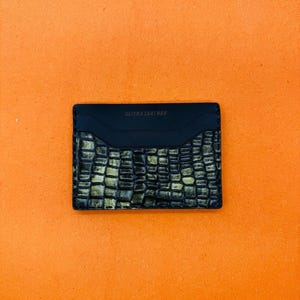 May include: A black leather card holder with a textured, crocodile-like pattern in shades of green and gold. The top section is smooth black leather, with the text "CLICKS LEATHER" embossed on it. The card holder is rectangular.