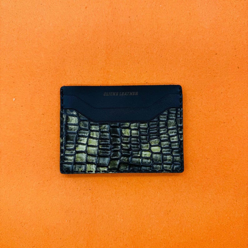 May include: A black leather card holder with a textured, crocodile-like pattern in shades of green and gold. The top section is smooth black leather, with the text "CLICKS LEATHER" embossed on it. The card holder is rectangular.
