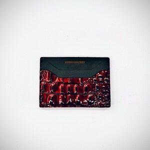 May include: A black and red card holder with a crocodile skin pattern. The top section is black leather with the text "CLICKS LEATHER" in gold lettering. The card holder is rectangular and sits on a white background.