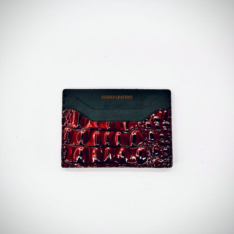 May include: A black and red card holder with a crocodile skin pattern. The top section is black leather with the text "CLICKS LEATHER" in gold lettering. The card holder is rectangular and sits on a white background.