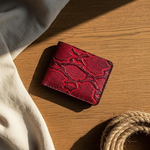 Op de afbeelding: Een rode bi-fold portemonnee met een textuur, slangenleer patroon. De portemonnee is gemaakt van leer en heeft een zwarte rand. De portemonnee ligt op een houten oppervlak met een touw en een witte doek.