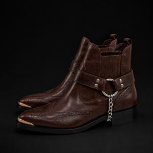 May include: A pair of brown leather ankle boots with a pointed toe and a decorative metal chain. The boots have a strap with silver hardware and a copper-colored metal accent on the toe. The boots are on a black background.