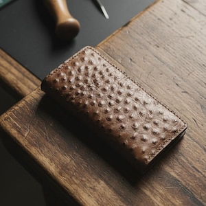 May include: A brown leather wallet with a textured, dotted surface rests on a wooden table. The wallet is rectangular and appears to be handmade. Leatherworking tools are visible in the background.