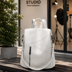 May include: White leather backpack with a top handle, shoulder straps, and a front zipper pocket. The bag has a curved design and is sitting on a wooden bench. The word "STUDIO PHOTOGRAPHY" is visible in the background.