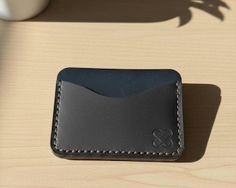 May include: A dark grey leather card wallet with a navy blue interior. The wallet has a stitched border and a debossed logo. The rectangular wallet is on a light wood surface. The wallet is approximately 10 cm long and 7 cm wide.