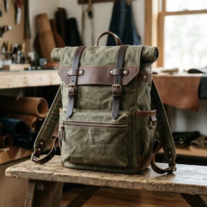 Può includere: Uno zaino roll-top in tela cerata verde oliva con cinghie in pelle marrone scuro, fibbie in ottone e una tasca frontale con cerniera. Lo zaino presenta spallacci regolabili, tasche laterali e finiture in pelle su tutto il design.